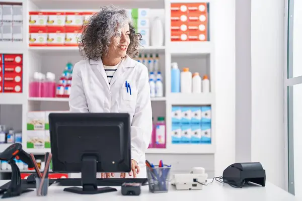 Middle age grey-haired woman pharmacist smiling confident using computer at pharmacy