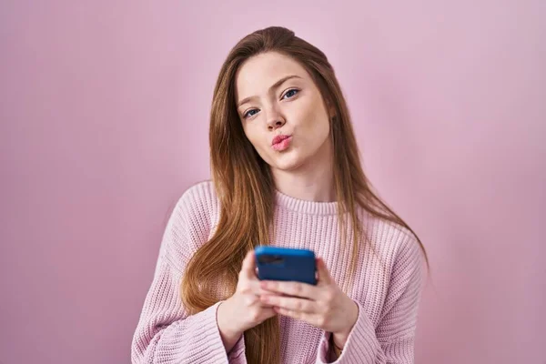 年轻的高加索女人用智能手机打字的信息看着摄像机吹一个吻是可爱的和性感的 爱的表达 — 图库照片