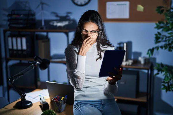 Young brazilian woman using touchpad at night working at the office smelling something stinky and disgusting, intolerable smell, holding breath with fingers on nose. bad smell