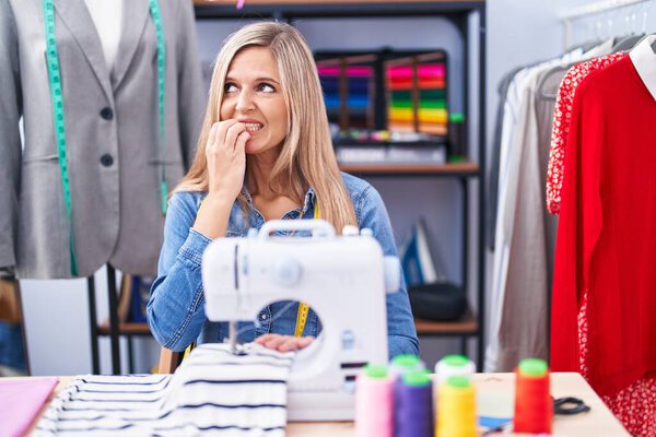 Blonde woman dressmaker designer using sew machine looking stressed and nervous with hands on mouth biting nails. anxiety problem. 