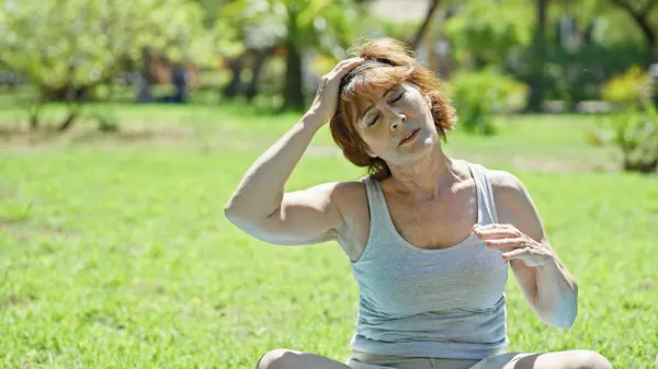 Middle age woman stretching head sitting on yoga mat at park