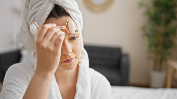 Young beautiful hispanic woman wearing bathrobe massaging face with skin gym at bedroom