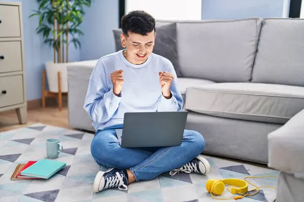 Non binary person studying using computer laptop sitting on the floor excited for success with arms raised and eyes closed celebrating victory smiling. winner concept.