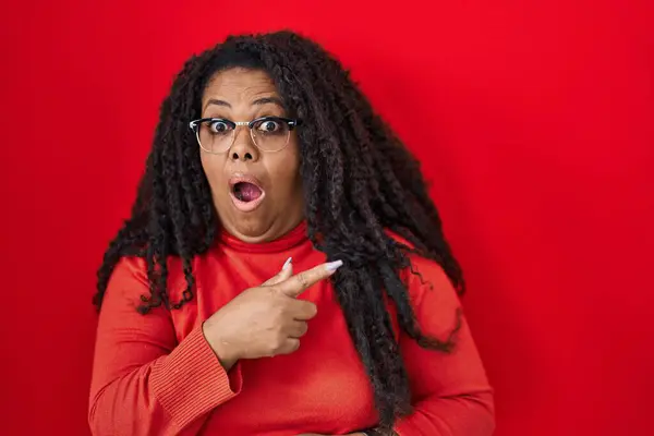 stock image Plus size hispanic woman standing over red background surprised pointing with finger to the side, open mouth amazed expression. 