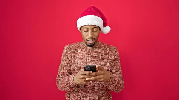 Afrikanischer Mann Trägt Weihnachtsmütze Mit Smartphone Über Isoliertem Rotem Hintergrund — Stockfoto