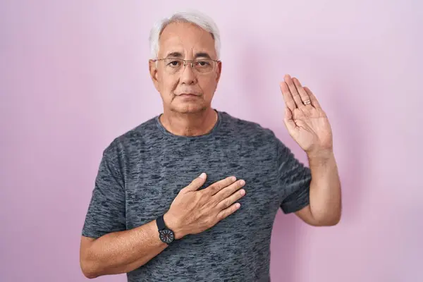 Middle age man with grey hair standing over pink background swearing with hand on chest and open palm, making a loyalty promise oath
