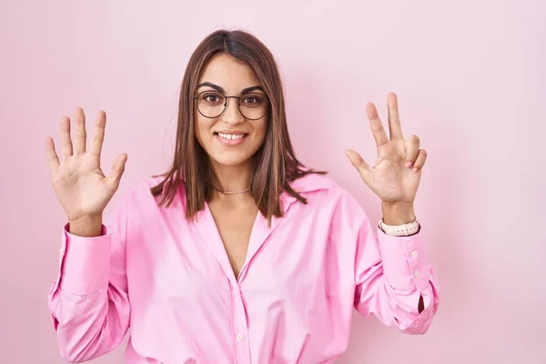 若いヒスパニックの女性は ピンクの背景の上に立って 自信と幸せに微笑んでいる間 指番号8を指で指で指で指を指さすことを身に着けています — ストック写真
