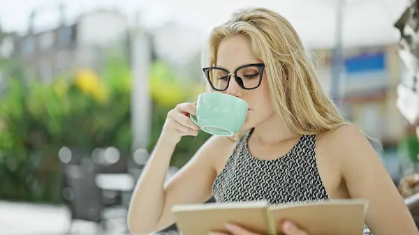 Young blonde woman drinking cup of coffee reading book at coffee shop terrace