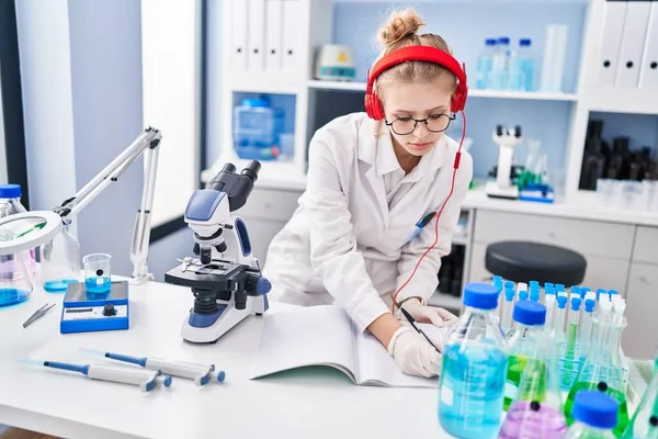 Young blonde woman scientist listening to music writing on notebook at laboratory