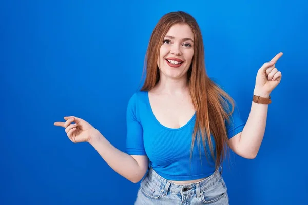 青い背景の上に立っている赤毛の女性は 異なる方向に指で指を指して自信を持って微笑んでいます 広告のためのコピースペース — ストック写真