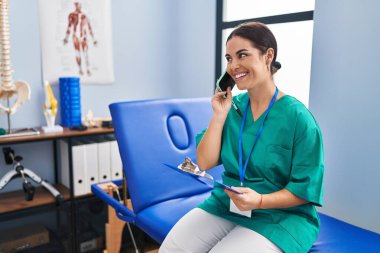 Young beautiful hispanic woman physiotherapist reading document talking on smartphone at rehab clinic