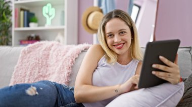 Young blonde woman using touchpad sitting on sofa at home