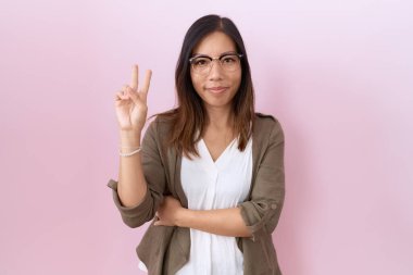 Orta yaşlı Çinli kadın pembe arka planda gözlük takıyor ve kameraya gülümsüyor zafer işareti yapıyor. İki numara.. 