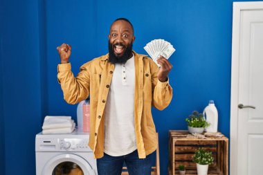 Afrikalı Amerikalı adam çamaşırhanede para tutuyor gururla bağırıyor, zaferi kutluyor ve başarıyı kollarını kaldırarak kutluyor. 