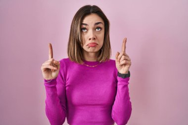 İspanyol kadın pembe arka planda dikiliyor üzgün ve üzgün görünüyor, parmaklarıyla yön gösteriyor, mutsuz ve depresif.. 