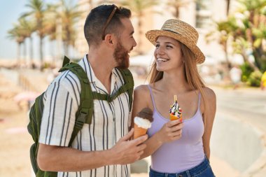 Kadın ve erkek turist, deniz kenarında dondurma yiyerek gülümsüyor.