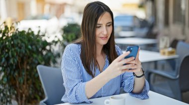 Akıllı telefon kullanan genç ve güzel bir İspanyol kadın kahve dükkanının terasında oturmuş gülümsüyor.