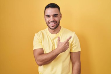 Young hispanic man standing over yellow background cheerful with a smile on face pointing with hand and finger up to the side with happy and natural expression 