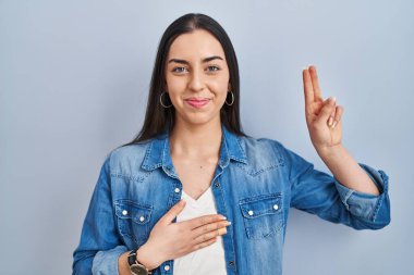 Hispanic woman standing over blue background smiling swearing with hand on chest and fingers up, making a loyalty promise oath 