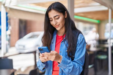 Latin kökenli genç kız sokakta akıllı telefon kullanarak gülümsüyor.