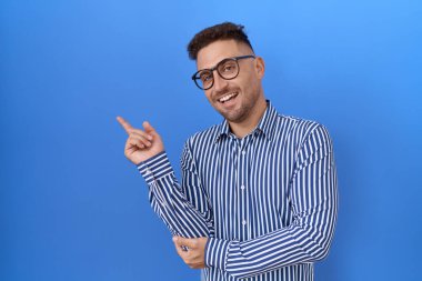 Sakallı, gözlüklü, yüzünde kocaman bir gülümseme olan, elini kameraya doğru gösteren İspanyol bir adam.. 