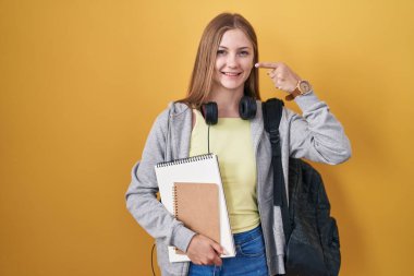 Young caucasian woman wearing student backpack and holding books pointing with hand finger to face and nose, smiling cheerful. beauty concept 