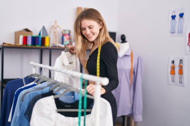 Young blonde woman tailor smiling confident holding clothes on rack at tailor shop