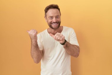 Sarı arka planda duran sakallı orta yaşlı bir adam. Arka tarafı işaret ediyor. Baş parmakları havada, kendinden emin gülümsüyor. 