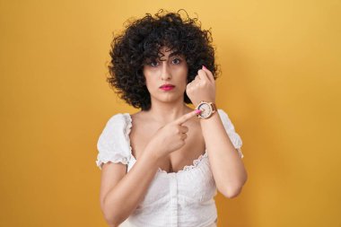 Sarı arka planda kıvırcık saçlı, genç esmer kadın. Saati, sabırsızlığı izlemek için acele ediyor. Rahat bir ifadeyle kameraya bakıyor. 