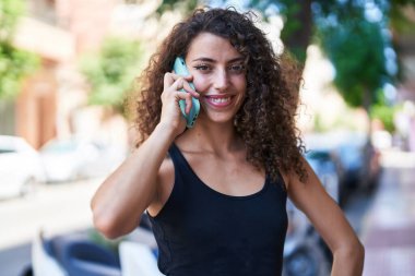 Genç, güzel, İspanyol bir kadın gülümsüyor ve caddede akıllı telefondan konuşuyor.
