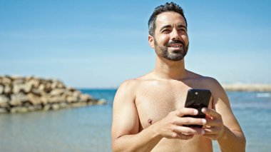 Genç İspanyol turist akıllı telefon kullanıyor plajda üstsüz duruyor.