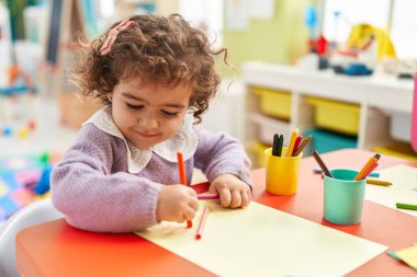 Adorable hispanic girl preschool student sitting on table drawing on paper at kindergarten