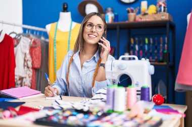 Young beautiful hispanic woman tailor talking on smartphone writing on notebook at clothing factory