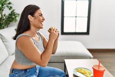 Young beautiful hispanic woman eating hamburger sitting on sofa at home