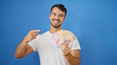 Genç İspanyol adam gülümsüyor. Meksika pesolarını soyutlanmış mavi arka planda tutuyor.