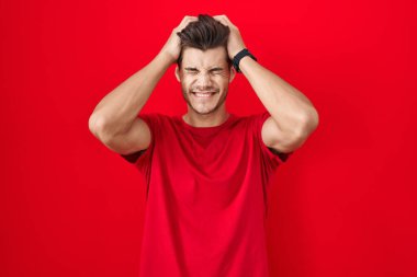 Genç İspanyol adam kırmızı arka planda duruyor. Baş ağrısı, acı ve migren yüzünden çaresiz ve stresli. Eller başının üstünde.. 