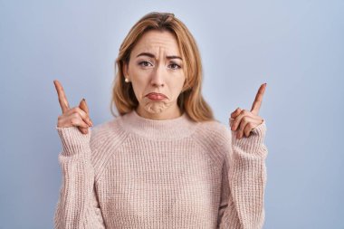 Hispanic woman standing over blue background pointing up looking sad and upset, indicating direction with fingers, unhappy and depressed. 