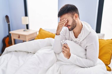 Young hispanic man taking pills for headache sitting on bed at bedroom