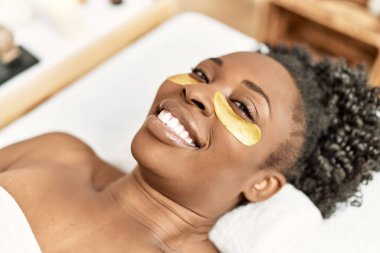 Masaj masasında uzanan Afrikalı Amerikalı kadın güzellik salonunda maske takıyor.