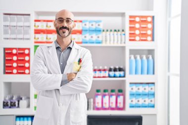 Young hispanic man pharmacist smiling confident standing with arms crossed gesture at pharmacy