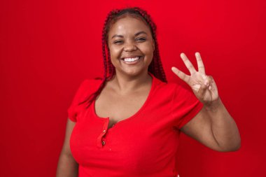 Kırmızı arka planda örülmüş saçları olan Afro-Amerikalı kadın üç numaralı parmaklarıyla gülümseyerek kendinden emin ve mutlu bir şekilde onu işaret ediyor.. 