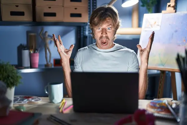 中年男人晚上坐在艺术工作室里 拿着笔记本电脑 兴奋地欢呼着 兴奋地举起双臂 睁开眼睛 庆祝着成功 获奖者概念 — 图库照片