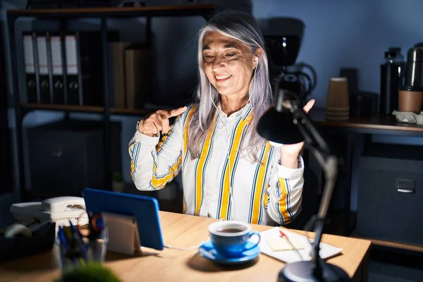 夜にオフィスで働く灰色の髪を持つ中年の女性は笑顔で 横に2本の手と指を指してカメラを見ています — ストック写真