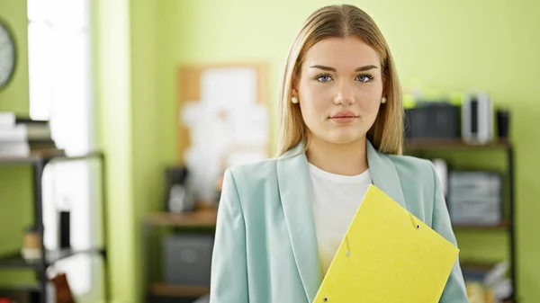Young blonde woman business worker holding binder with serious face at office