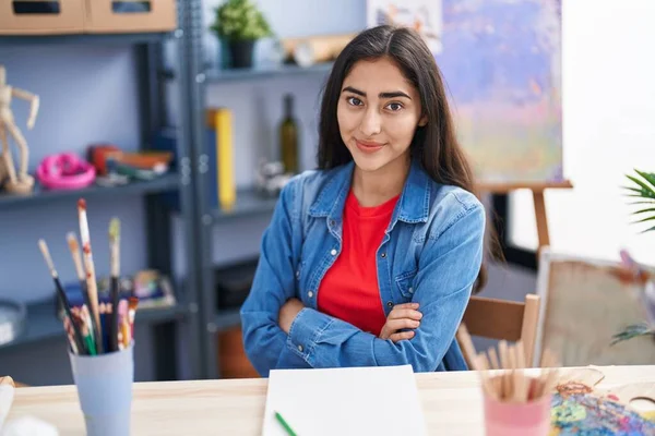 stock image Young hispanic girl artist sitting on desk with arms crossed gesture at art studio