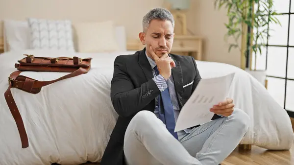 Young hispanic man business worker reading document sitting on floor thinking at hotel room