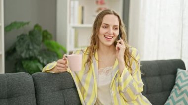 Young blonde woman talking on smartphone drinking coffee at home