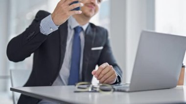 Young hispanic man business worker using laptop talking on smartphone at office