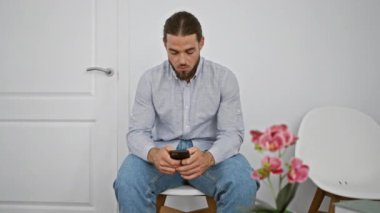 Genç İspanyol adam akıllı telefon kullanıyor. Sandalyeye oturmuş, bekleme odasına bakıyor.