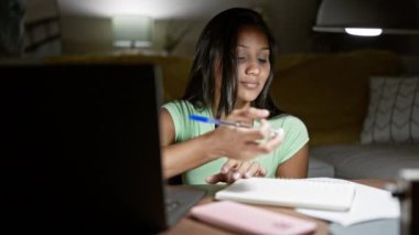Laptop kullanan genç Latin kadın evde not alıyor.